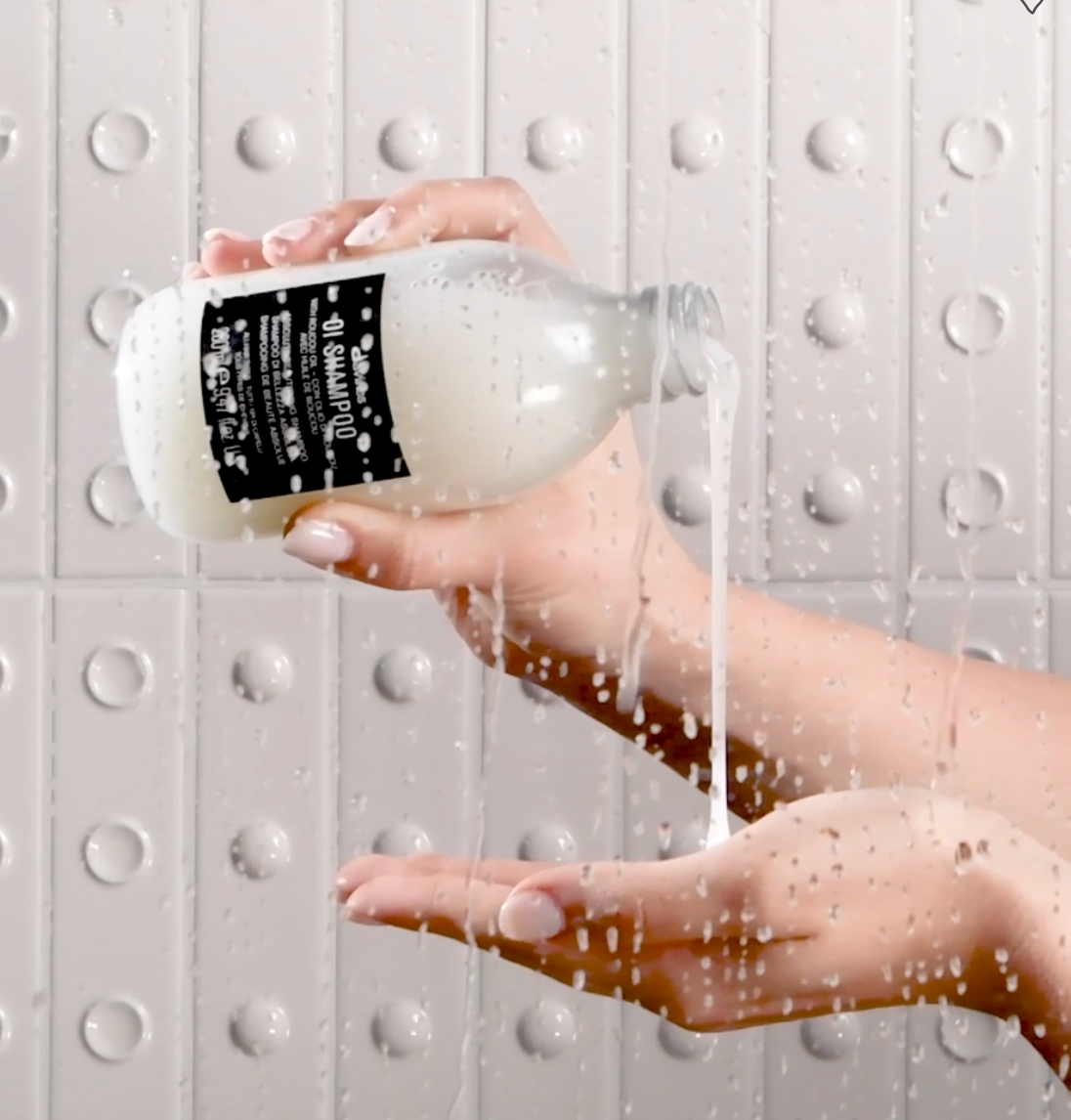 Hand holding a bottle of shampoo with water flowing, against a textured gray background. Natural hair care products.